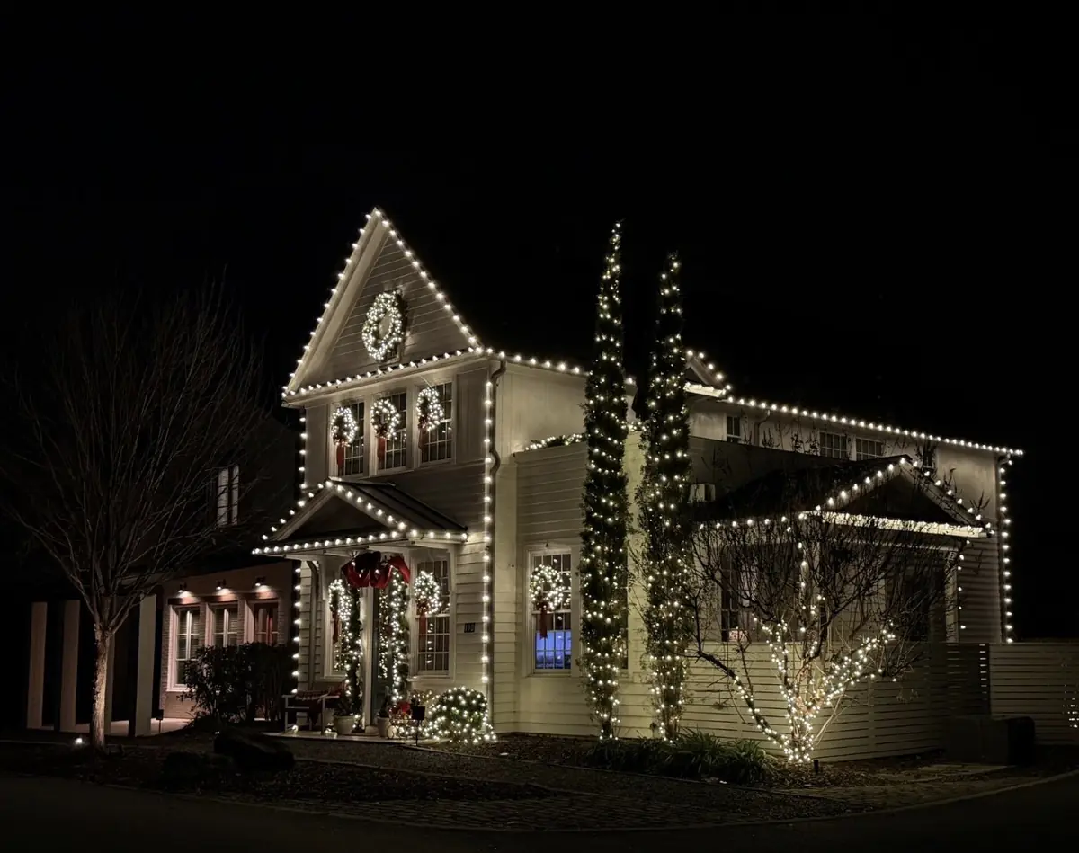 White two-story home outlined in warm white Christmas lights with lit wreaths on every window and wrapped trees, installed by Bassett Bro Holiday Lighting